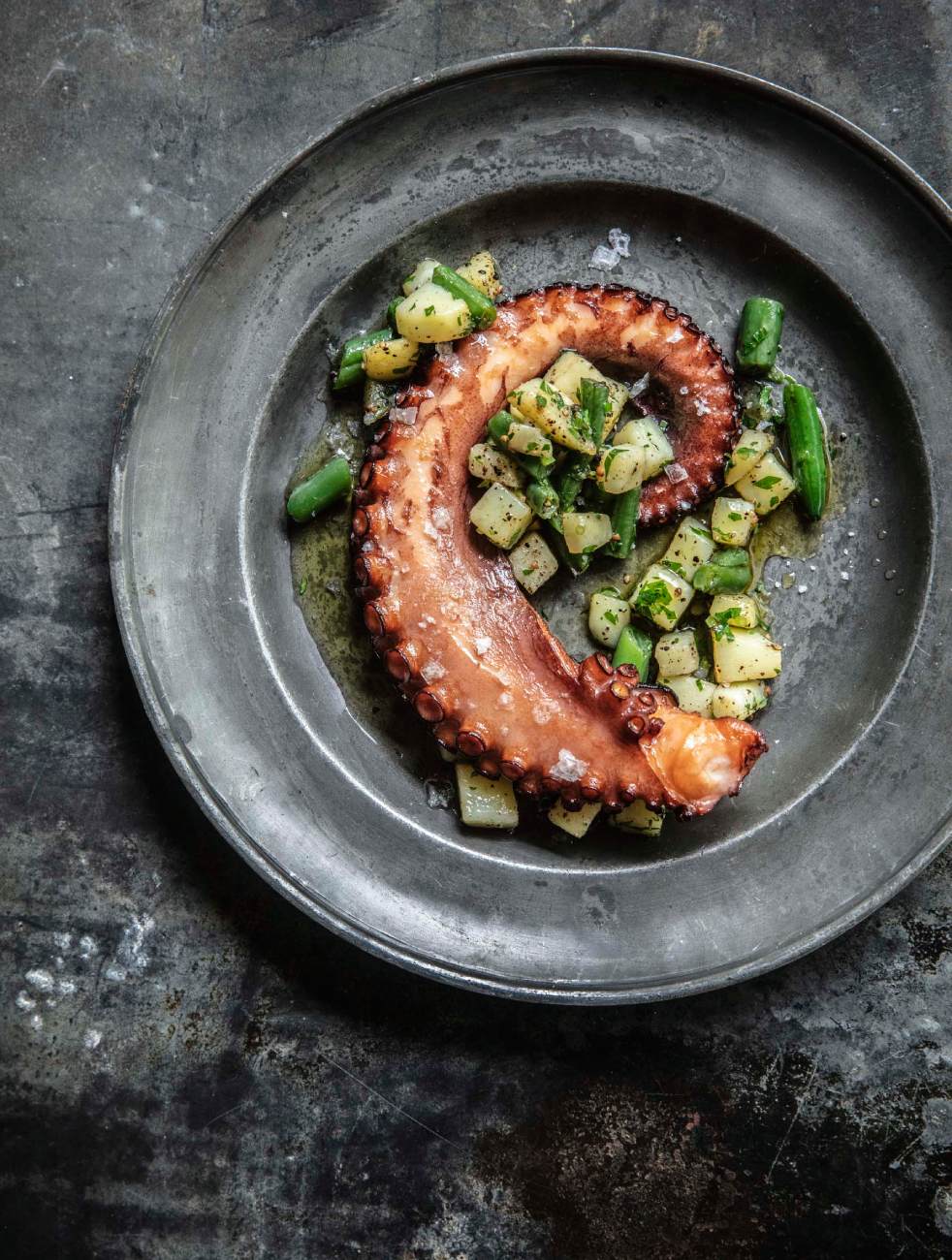 Overhead shot of grilled octopus tentacle with potatoes and green beans on an aged, textured metal plate and background.