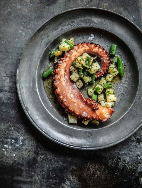 Overhead shot of grilled octopus tentacle with potatoes and green beans on an aged, textured metal plate and background.
