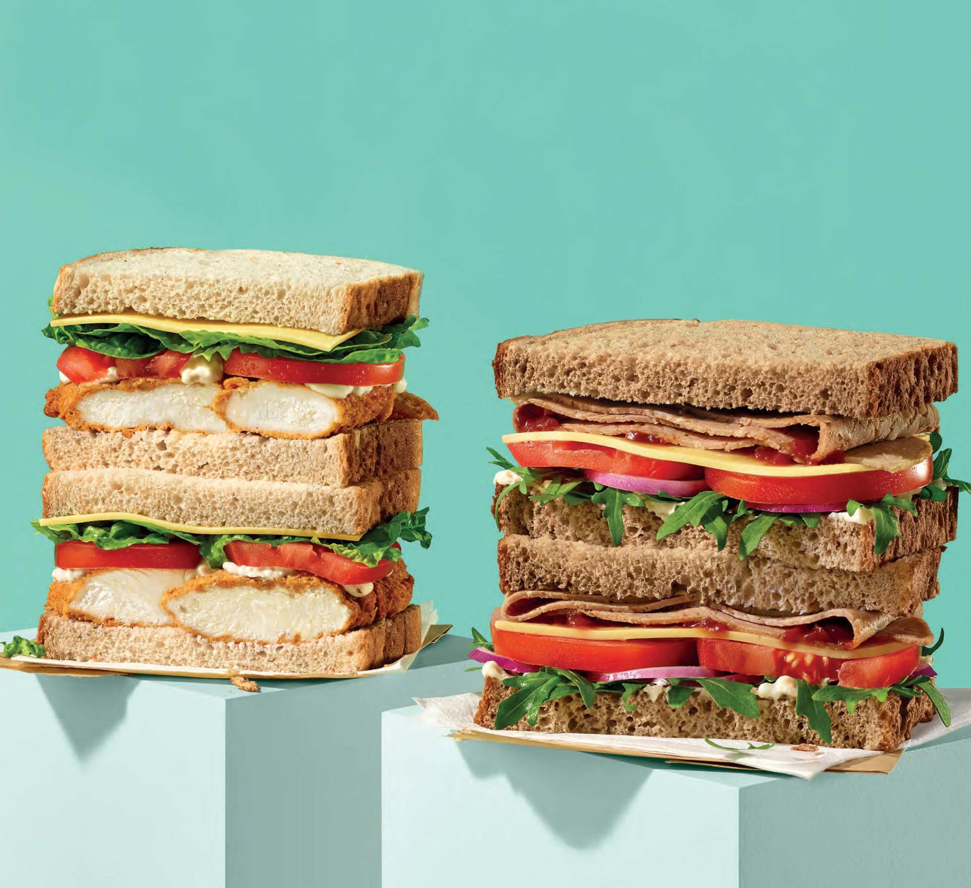 Two stacked sandwiches with chicken, cheese, tomatoes, and lettuce on a light teal background, food photography.