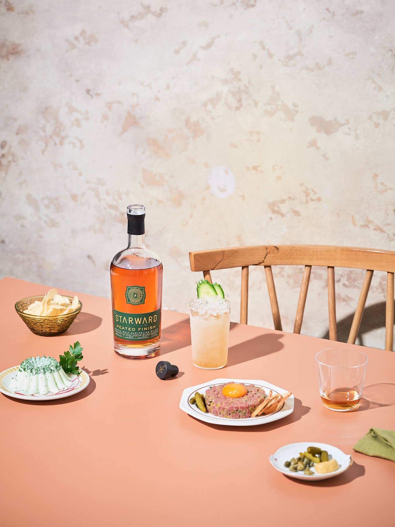 Still life shot of a bottle of Starward whisky, cocktails, and plates of food, displayed on a peach-colored table against a textured wall.