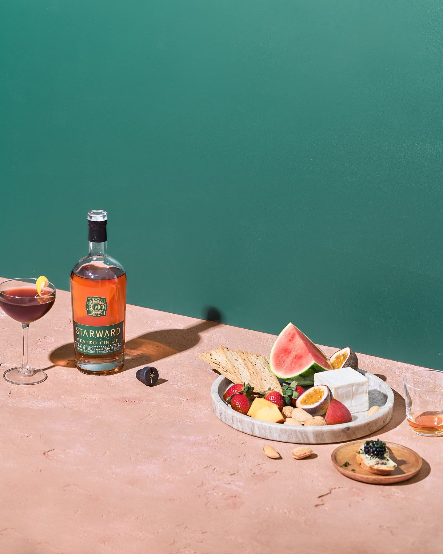 Still life shot of a Starward whisky bottle beside a cocktail, a cheese board with watermelon and other fruit, against a teal backdrop.