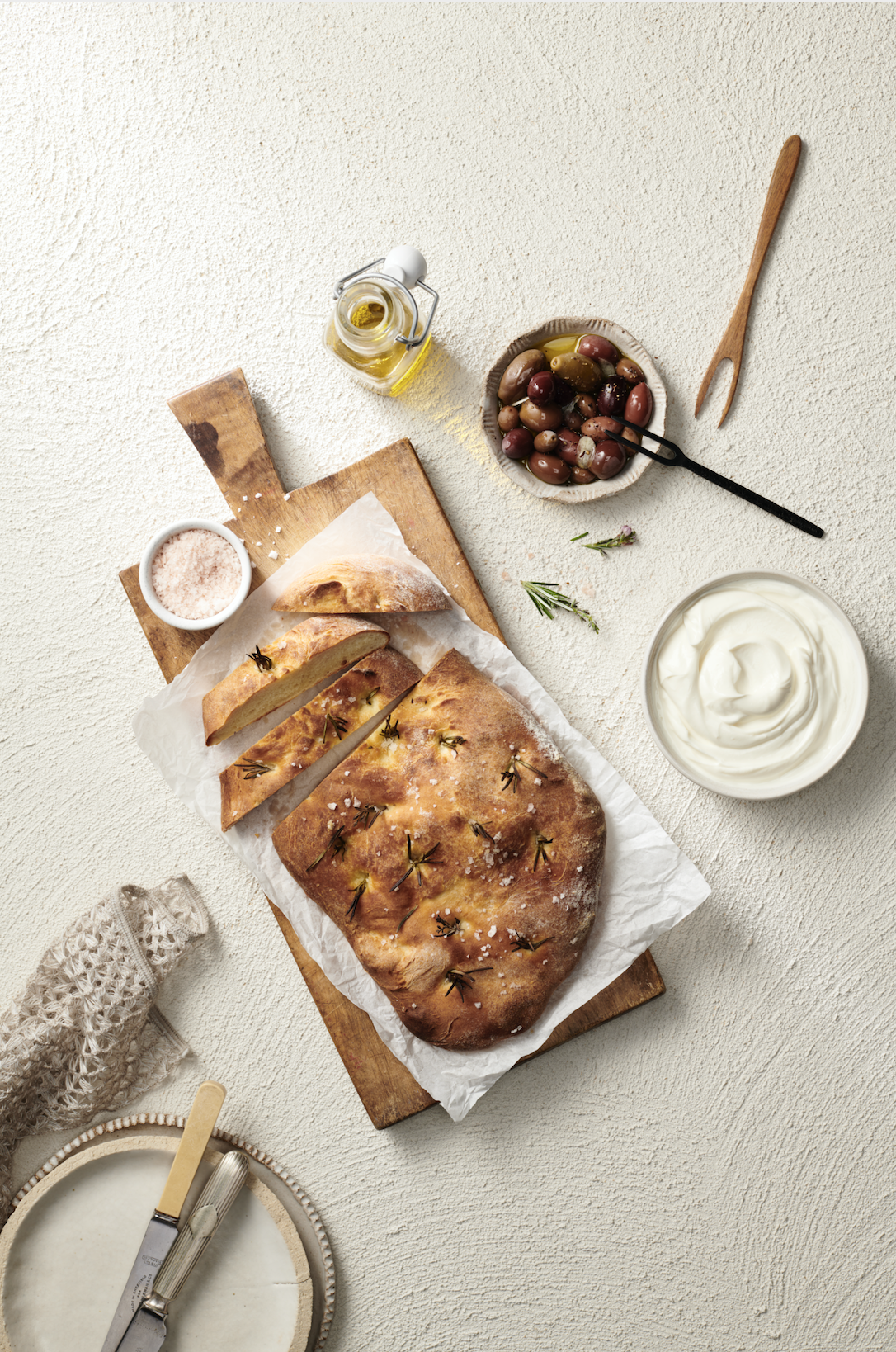Overhead shot of focaccia bread on a wooden cutting board with olive oil, olives, sea salt, and yogurt on a white textured surface.