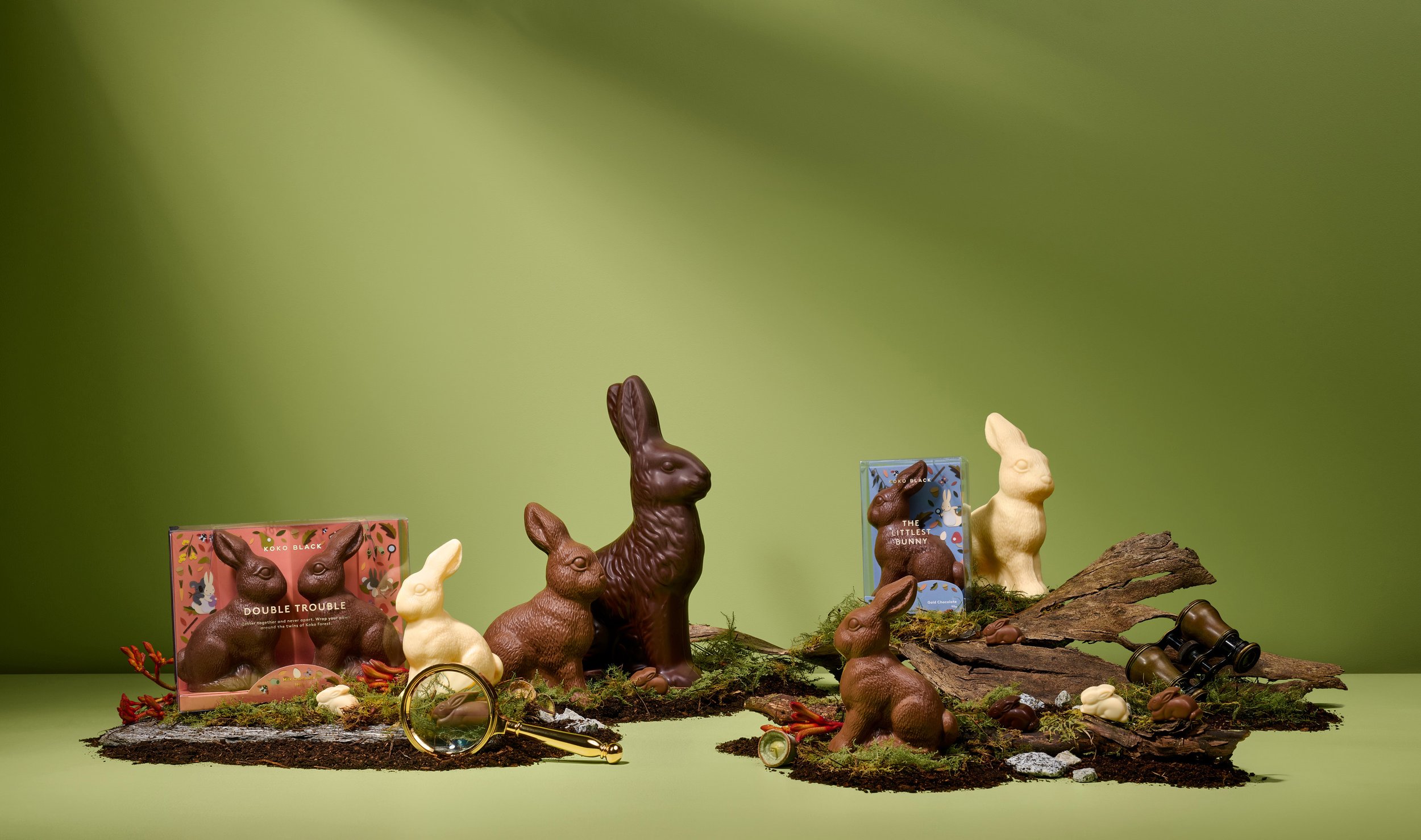 Chocolate Easter bunnies, two boxes, magnifying glass, and binoculars arranged on moss against a green backdrop.