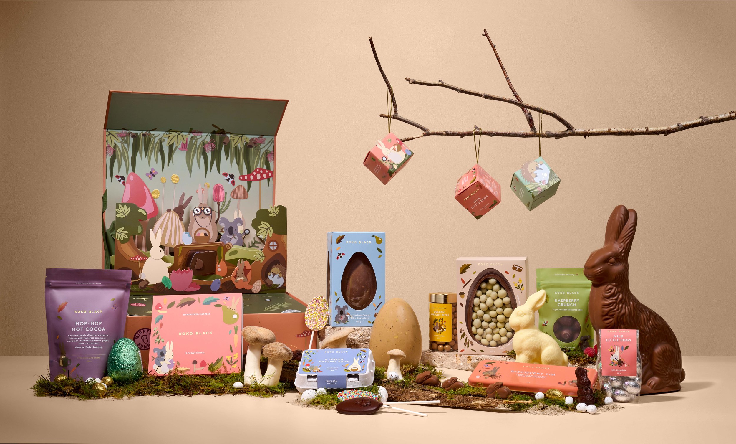Cocoa-themed Easter product arrangement featuring chocolate eggs, bunnies, and woodland scene packaging on a soft tan backdrop.