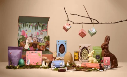 An array of Easter chocolate products, gift boxes, and decorative bunnies arranged on moss against a light background with hanging treat boxes.