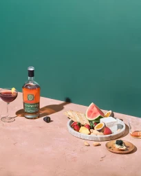 Bottle of whisky, a cocktail, and a marble platter with fruit, cheese, and crackers styled on a pink surface.