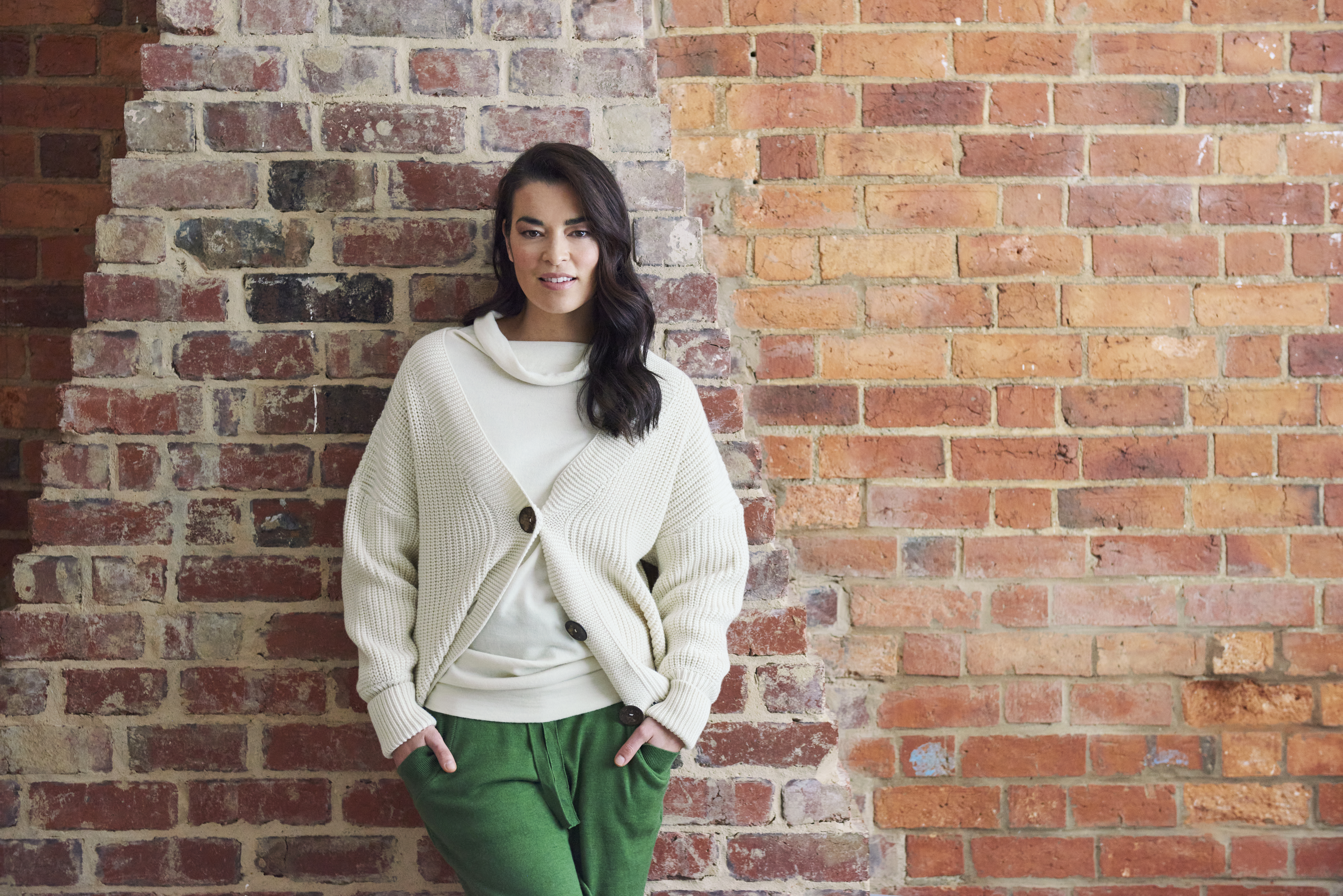A woman with dark hair poses in front of a rustic brick wall, wearing a cream-colored sweater and green pants, in a relaxed indoor setting.