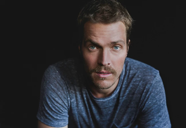 Portrait of a man with short brown hair and blue eyes wearing a textured blue t shirt against a dark background