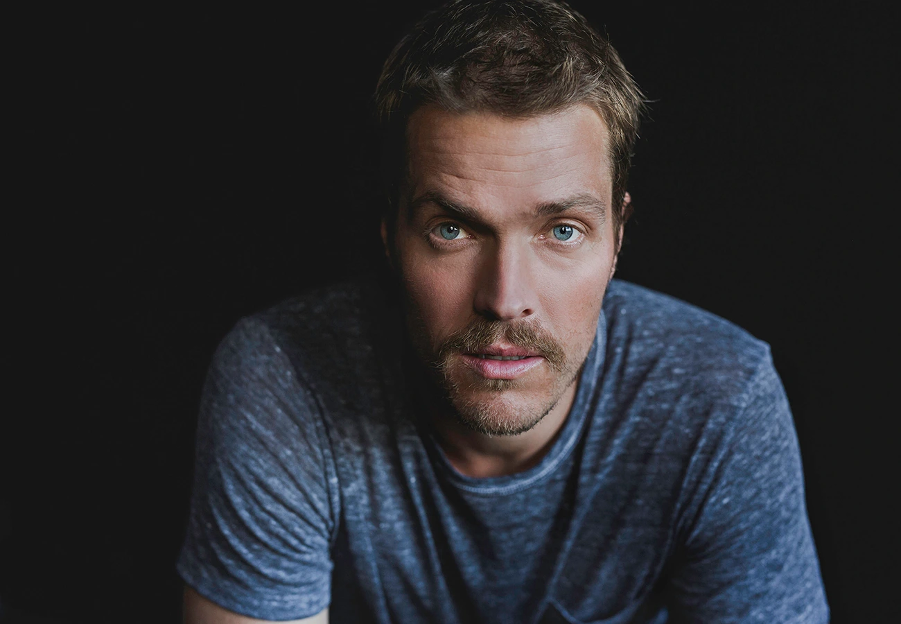 Portrait of a man with short brown hair and blue eyes wearing a textured blue t shirt against a dark background