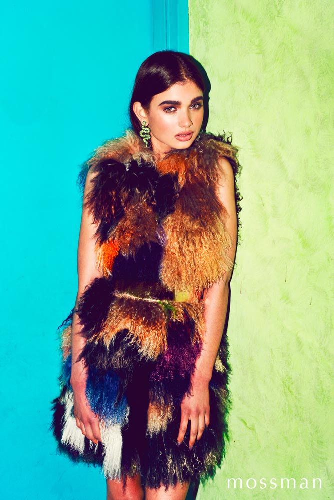 Fashion shot of a woman with dark hair wearing a multi-colored fur vest and large green earrings, standing against a bright blue and green wall.