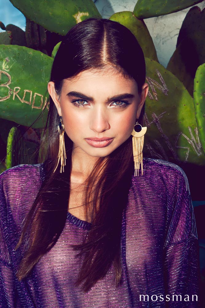 Eye-level portrait of a young woman with dark hair and gold earrings wearing a purple shirt in front of a green cactus plant.