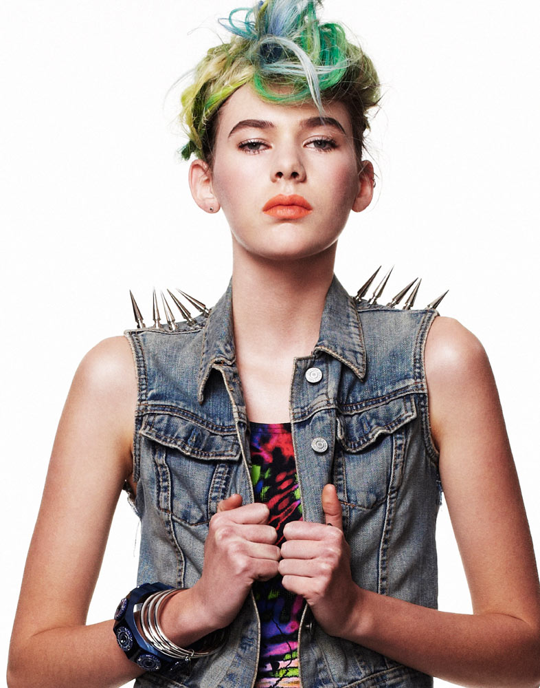 Stylish young woman with vibrant dyed hair poses indoors wearing a spiked denim vest and colorful top against a bright white backdrop.