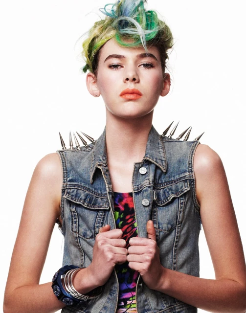 Stylish young woman with vibrant dyed hair poses indoors wearing a spiked denim vest and colorful top against a bright white backdrop.
