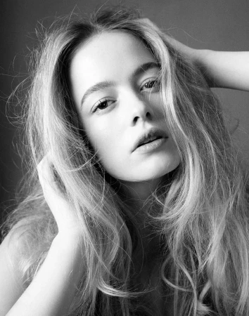 Close-up black and white portrait of a young woman with tousled long blonde hair posing with her hands near her face