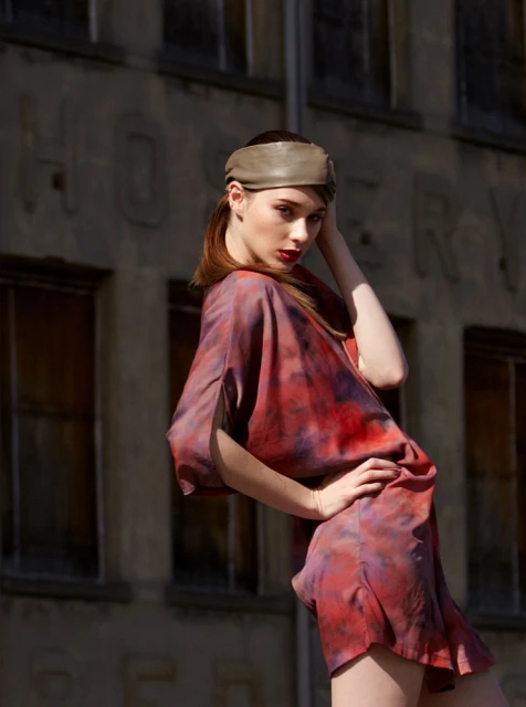 A young woman in a tie-dye mini-dress poses in front of a weathered building with large windows and faded signage in an urban setting.