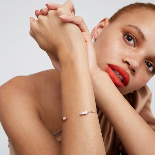 A person with neutral expression wearing a rose-gold bracelet, bright red lipstick, and a short haircut is posing in a close-up shot.