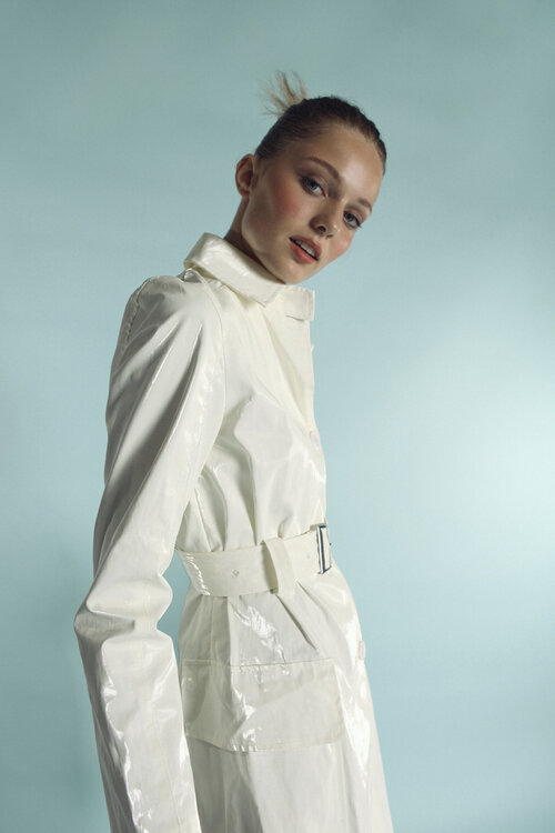 Portrait of a young female model in a shiny white belted coat against a light blue background in a studio fashion shot.
