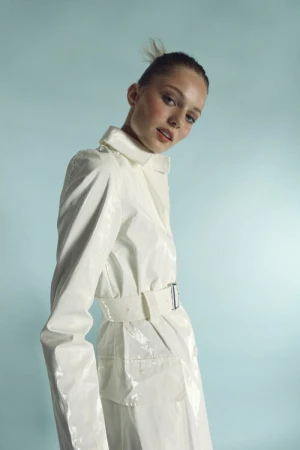 Model in a glossy white belted coat with hair styled in an updo against a light blue seamless studio background.