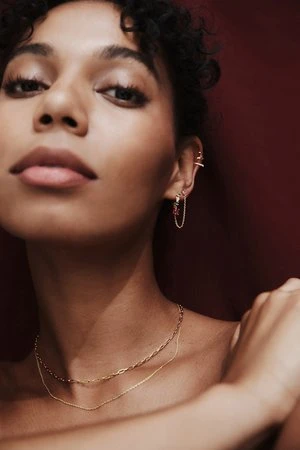 A close-up studio shot of a woman with dark hair and gold jewelry, including earrings and a necklace, against a dark red background.