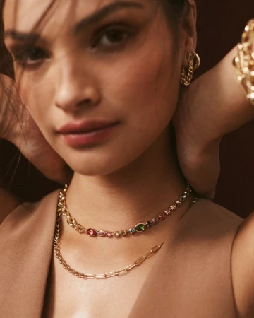 Close-up portrait of a woman wearing gold chain necklaces with colourful gemstones and hoop earrings.