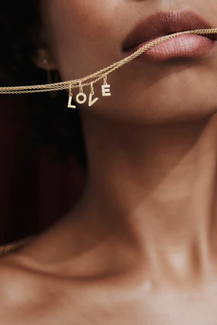 Close-up of a person's face holding a delicate gold necklace with 'LOVE' letter charms in their mouth, showcasing fashion and beauty.
