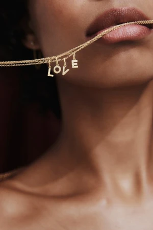 Close-up of a person's face holding a delicate gold necklace with 'LOVE' letter charms in their mouth, showcasing fashion and beauty.