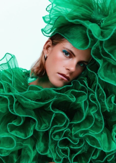 A woman with short blonde hair, adorned in a ruffled green tulle garment and matching headpiece, is shown against a plain white background.