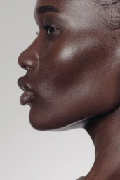 Close-up profile portrait of a black woman with smooth skin and subtle lip gloss against a neutral grey studio background