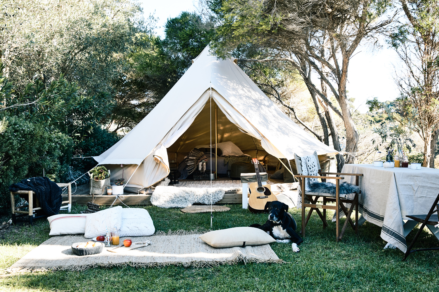 Canvas bell tent setup on green grass, a dog lounging on a rug, surrounded by outdoor seating and trees in a backyard.