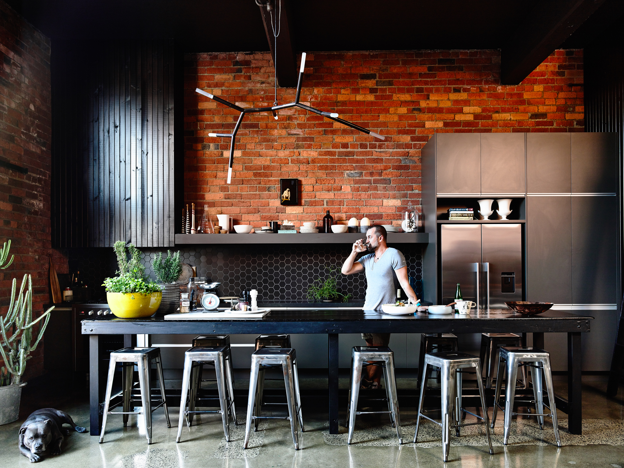 Modern kitchen interior featuring a man drinking water at a long black table with stools, brick wall, and a dog lying on the floor.