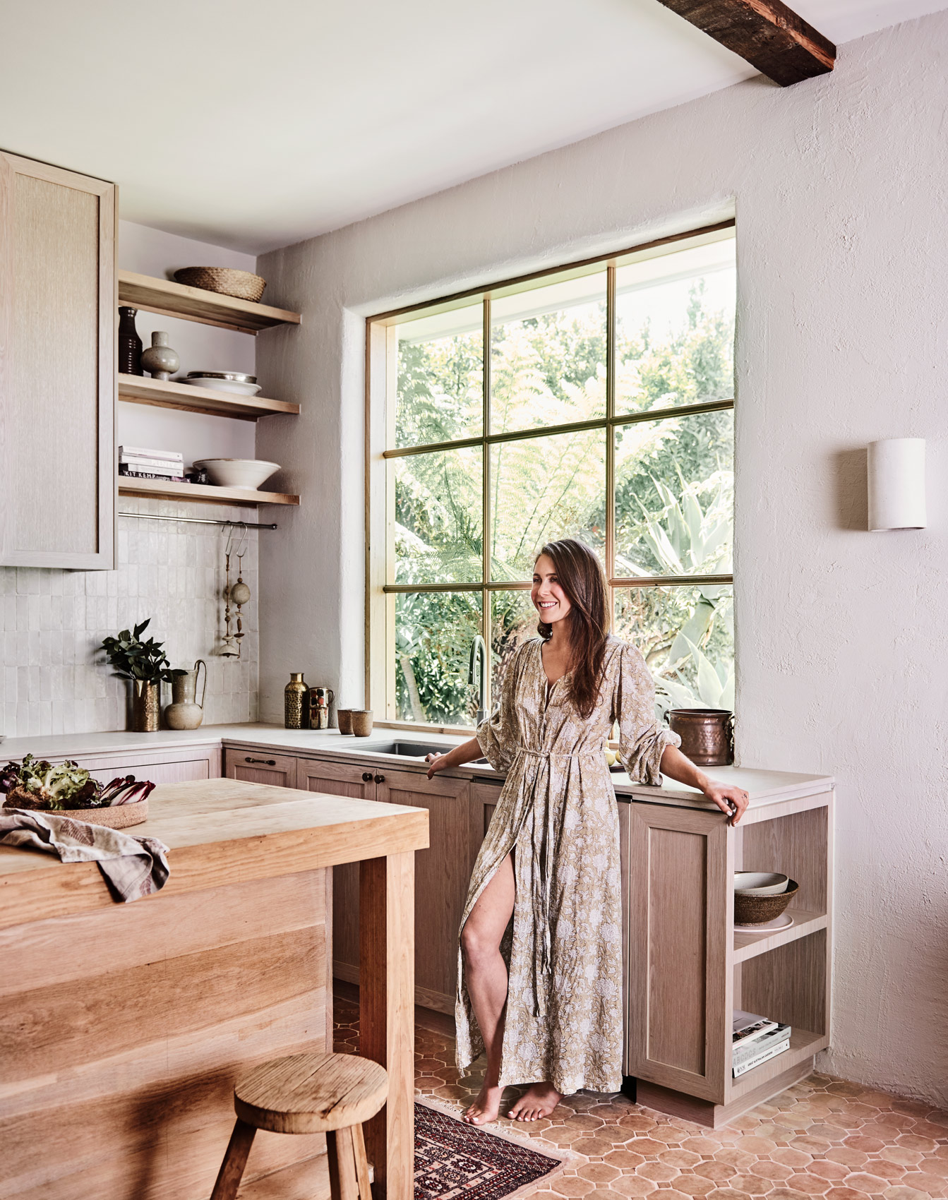 Woman in long dress stands barefoot in sunlit neutral-toned kitchen with wooden island, stool, open shelves, and large window overlooking foliage.
