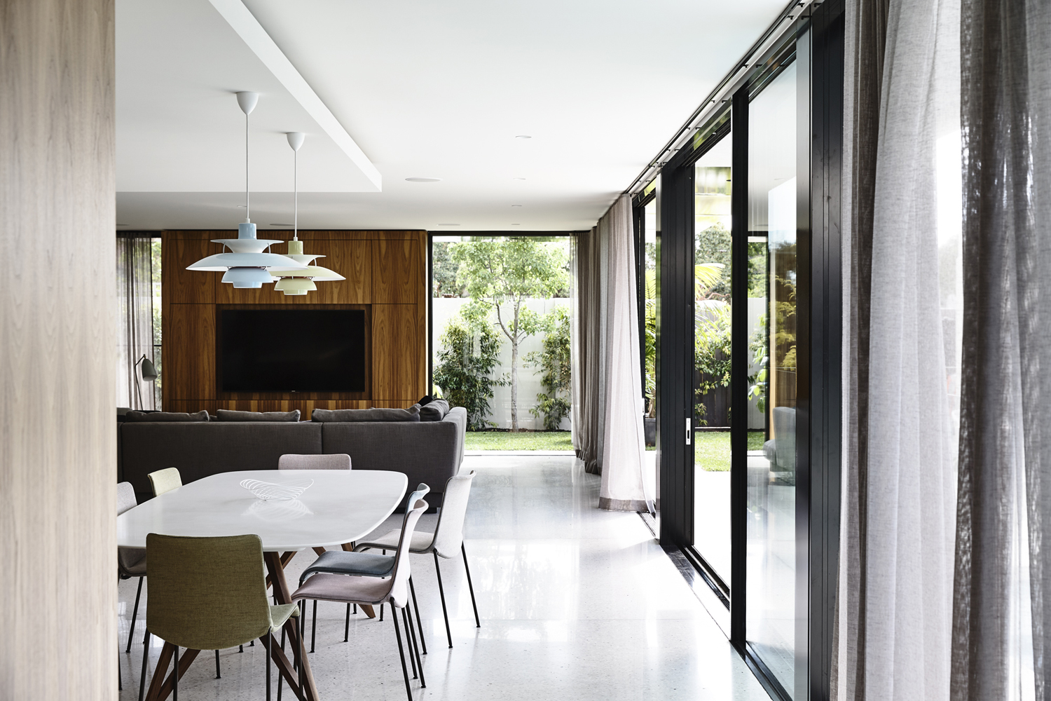 Contemporary interior with dining table, chairs, and modern pendant lights. A living area with a grey couch and a view of the garden.