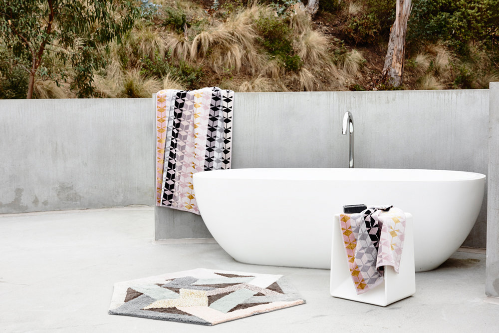 Modern bathroom interior featuring a white freestanding bathtub, patterned towels, geometric rug, and minimalist decor near a window.