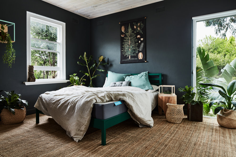 Bedroom interior with a green bed frame, houseplants, a jute rug, and a graphic wall hanging in a natural light-filled space.