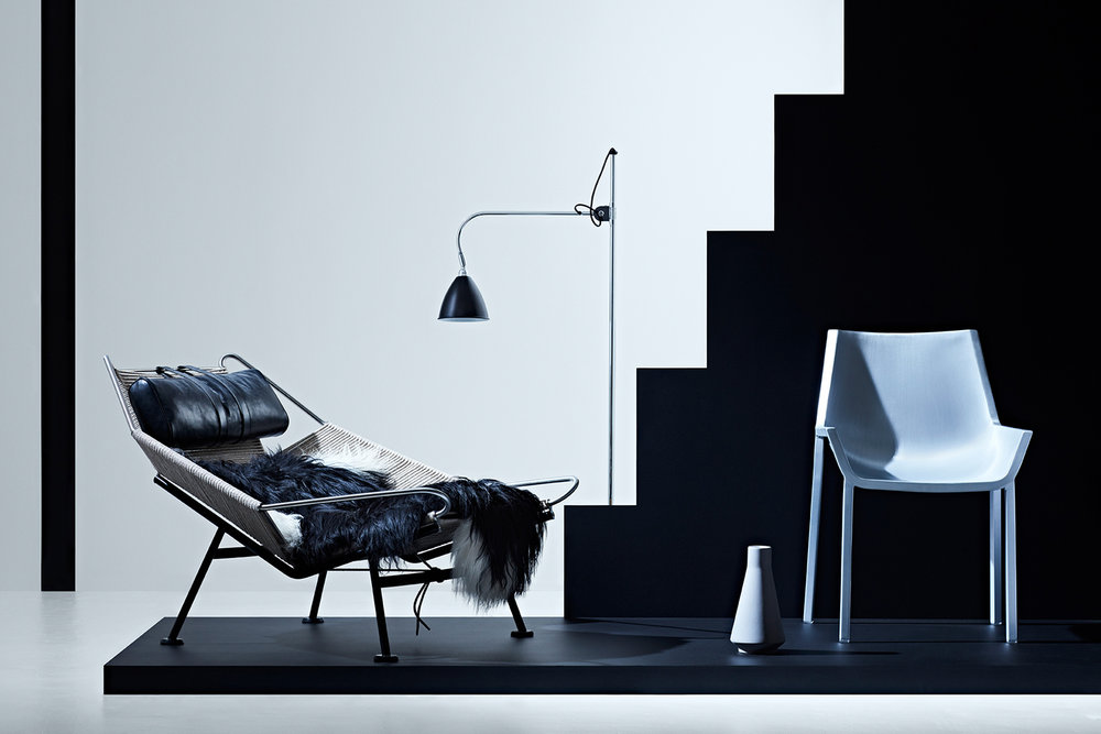 Interior photo of a modern lounge chair with black cushion, white chair, lamp, and vase on a black and white background.