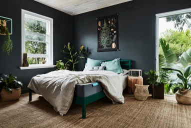 Modern bedroom with dark grey walls, a green wooden bed frame, linen bedding, and various potted indoor plants by Heather Nette King