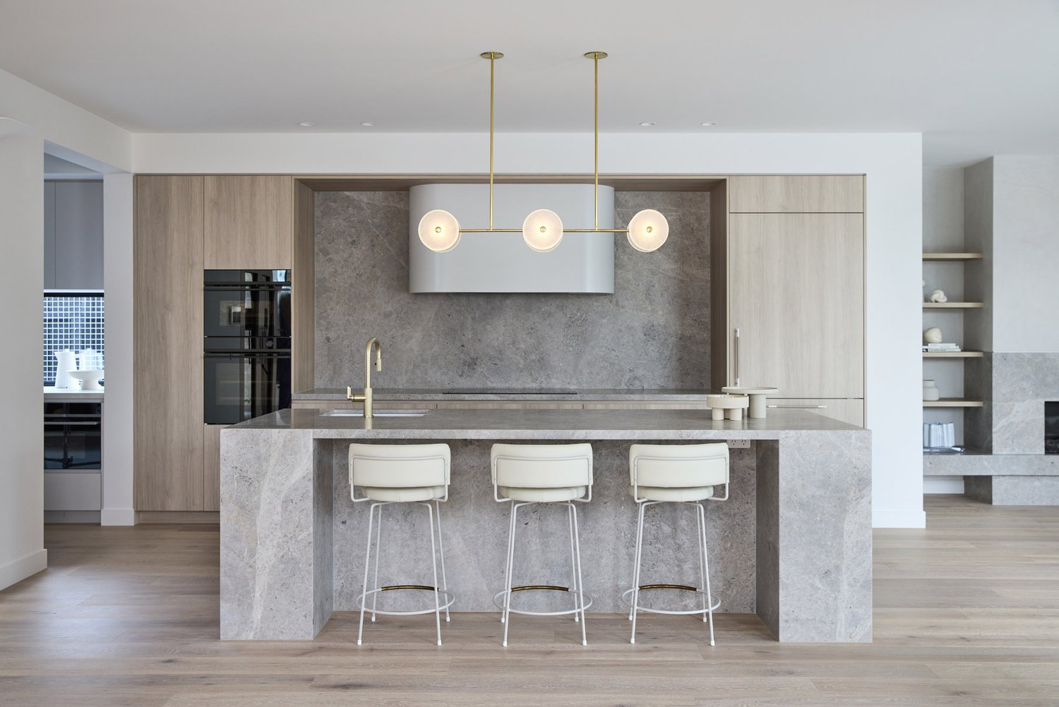 Modern kitchen interior featuring a marble island with three stools, a contemporary chandelier, and light wood cabinetry.