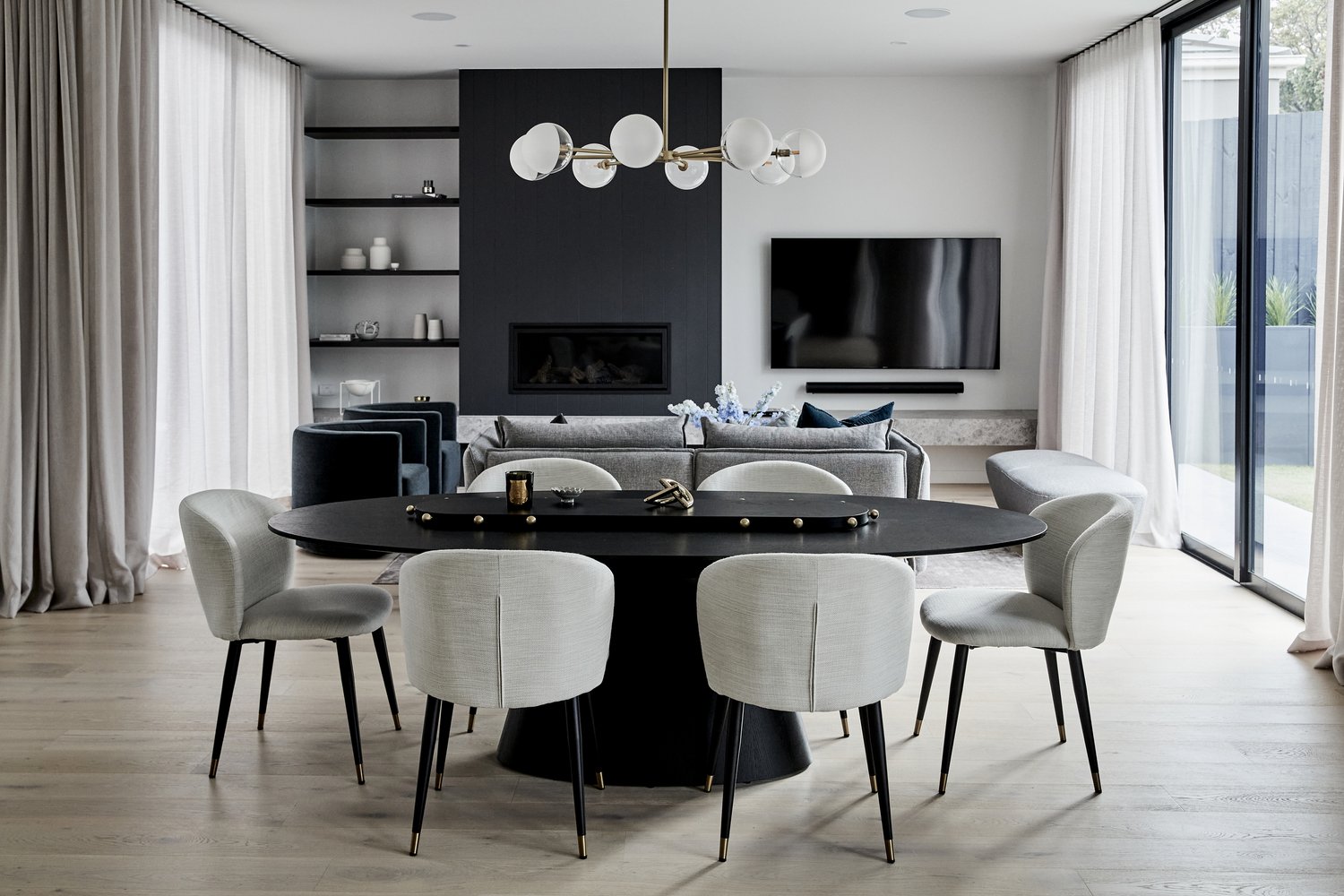Interior view of a modern dining room with a black oval table surrounded by six grey chairs, a chandelier, and a view to the backyard.