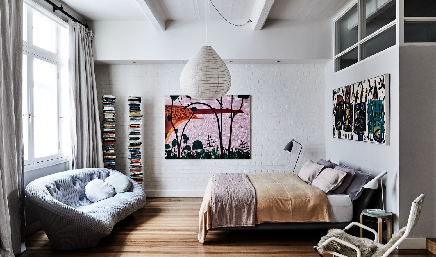Bedroom interior with bed, grey couch, modern art, white brick walls, and wood floors, creating a cozy and contemporary space.