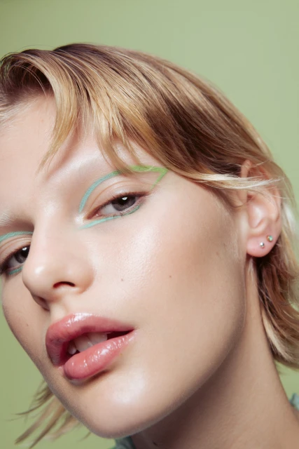 Close-up portrait of a fair-skinned young adult woman with blonde hair, light green eyeshadow, and glossy lips against a light green backdrop.