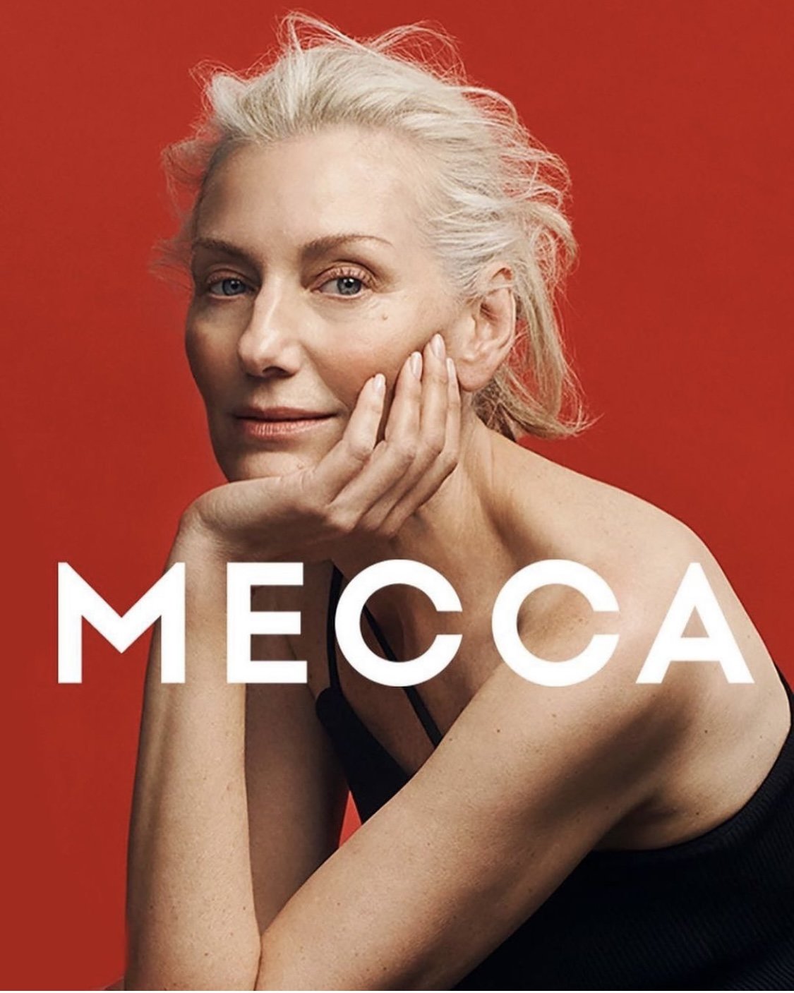 Close-up portrait of an elderly woman with light skin and short white hair in a black tank top, resting her face on her hand.