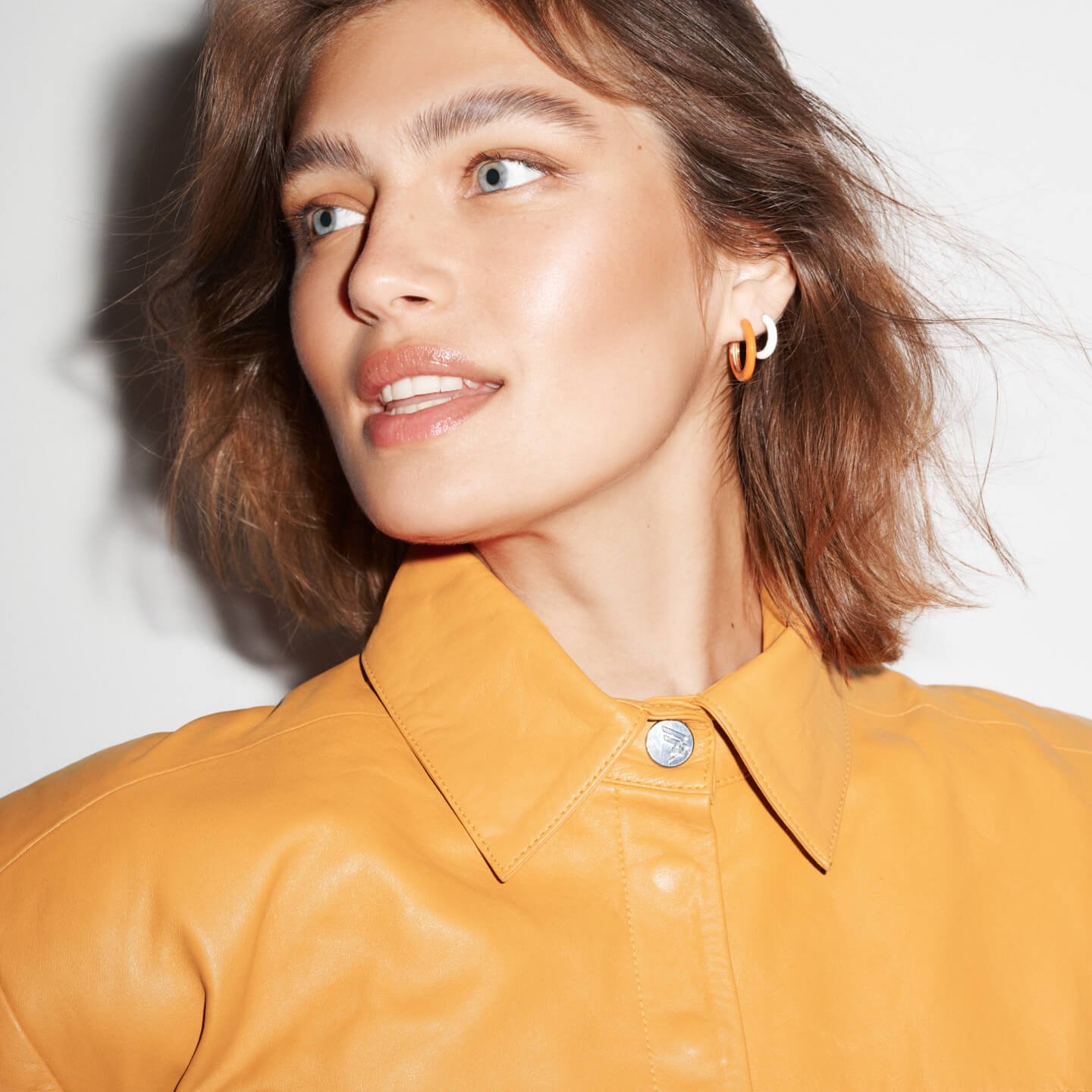 Close-up portrait of a woman with fair skin, blue eyes, and light brown hair, wearing a yellow leather jacket and two-tone hoop earrings.