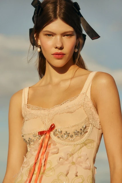 Close-up of a young woman with black ribbons in her hair, wearing a pale pink floral dress against a light blue sky backdrop.