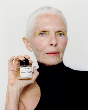 Close-up portrait of an elderly woman holding a perfume bottle with yellow eyeshadow against a white seamless background