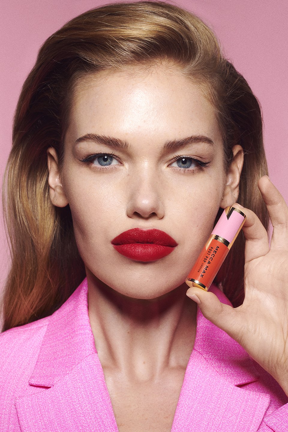 Close-up of a blond woman holding a coral-colored lip gloss tube, wearing a pink blazer against a pink background in studio lighting.