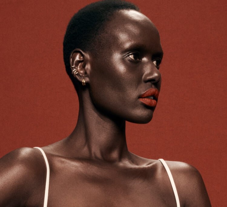 Close-up studio shot of a Black woman with short hair and red lipstick, wearing a white tank top, against a warm reddish-brown background.