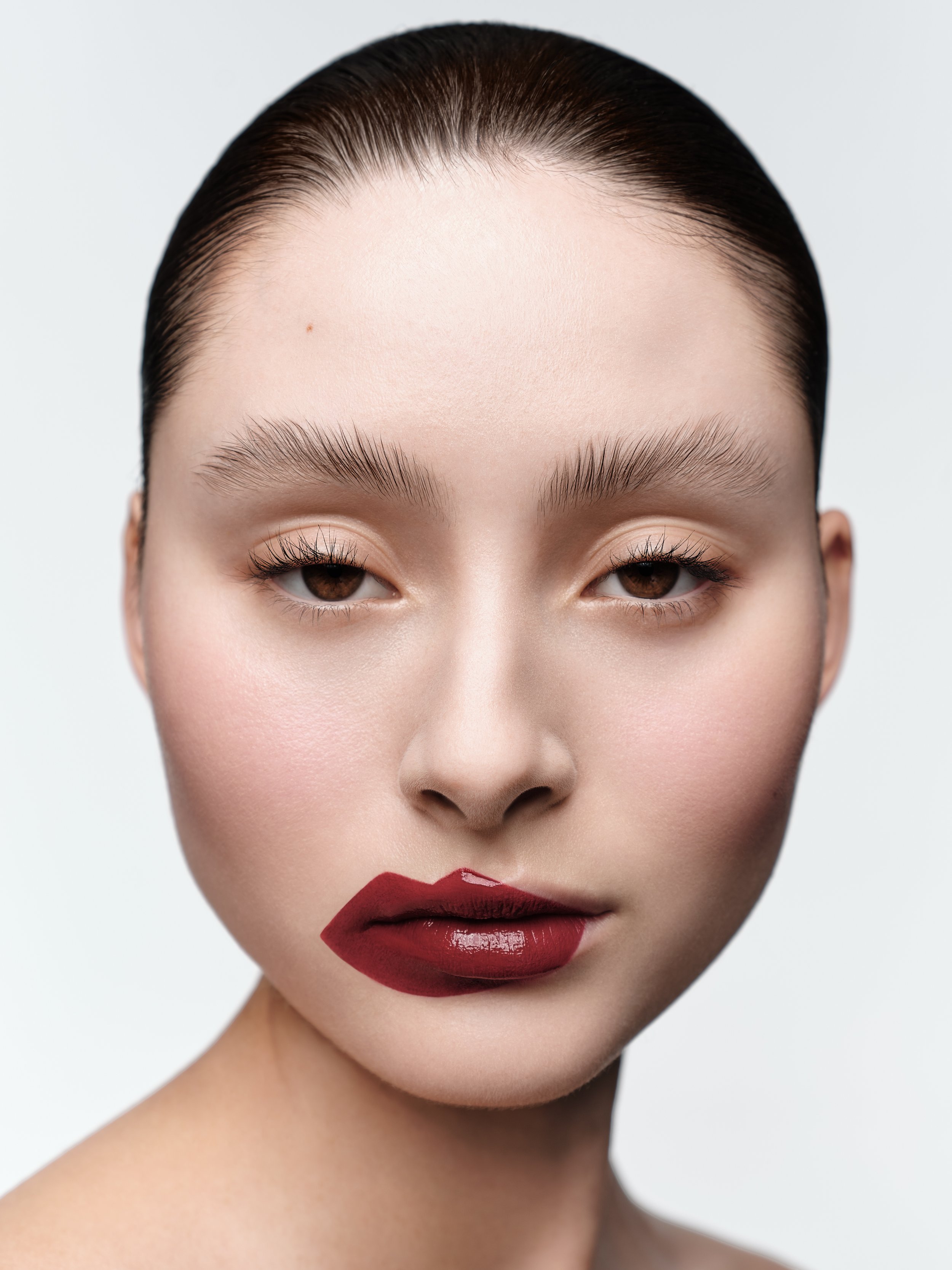 Close-up beauty portrait of a woman with burgundy lipstick partially covering her face, set against a clean, light background.
