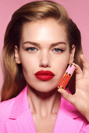 Close-up portrait of a woman with bold red lipstick holding a pink lipstick tube, wearing a pink blazer against a pink backdrop