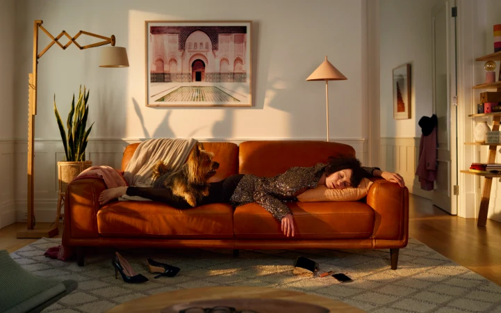 A woman and a dog rest on a couch in a living room, with scattered accessories nearby, and a framed picture hanging on the wall.