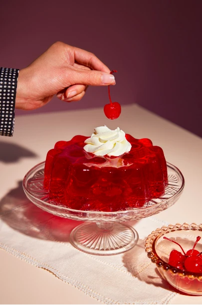 A hand places a cherry on top of a red gelatin dessert with whipped cream, set on a glass cake stand with a pink background.
